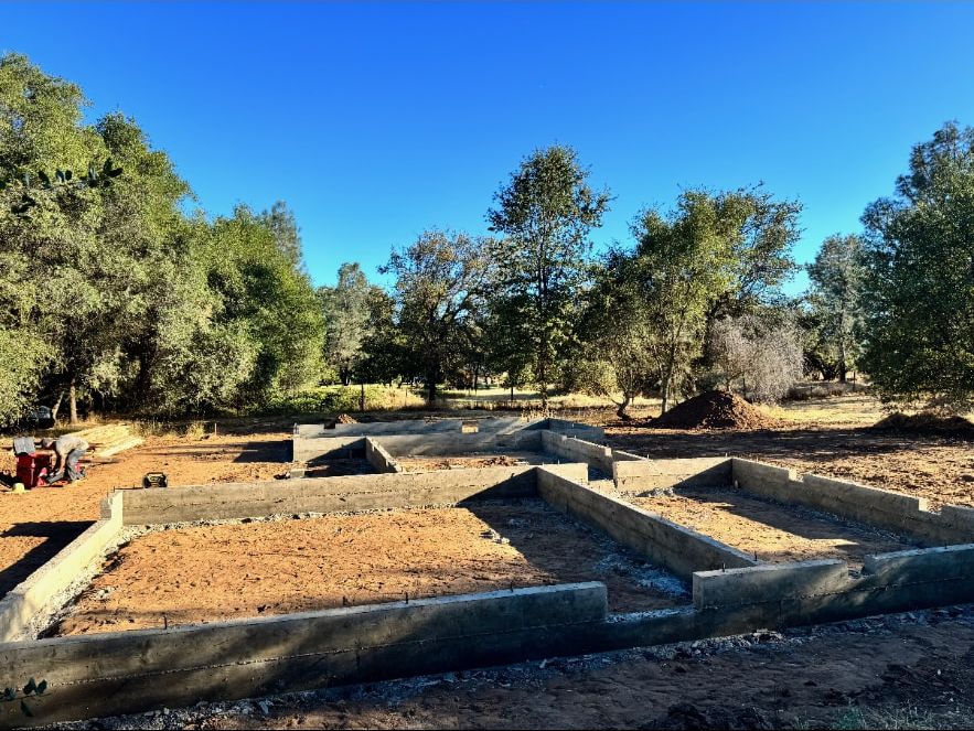 Concrete foundation walls laid out across a graded building pad surrounded by valley oaks